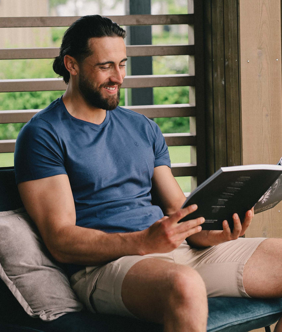 Man met blauw T-shirt met ronde hals en UM (undiemeister) logo en beige broek zit op een bankje een tijdschrift te lezen