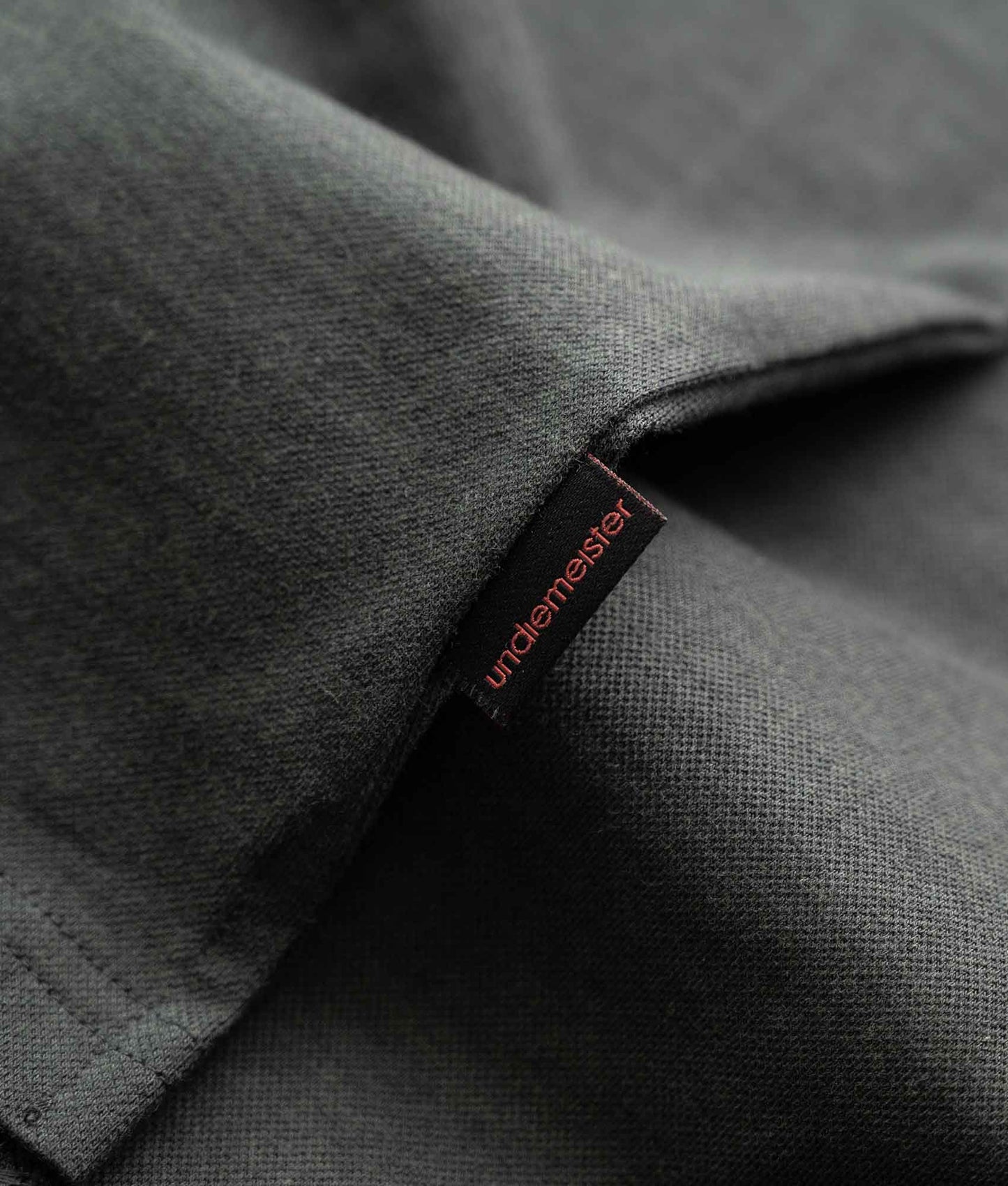 Close-up van grijze polo met zwart label met daarop in rode letters het Undiemeister logo